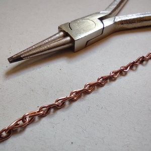 Peut inclure: Gros plan sur une pince argentée avec une chaîne en cuivre à côté sur une surface blanche.