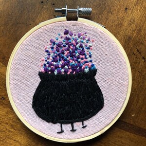 May include: Embroidered hoop art featuring a black cauldron with pink, purple, and blue confetti spilling out of it. The cauldron has small legs and is on a pink background.