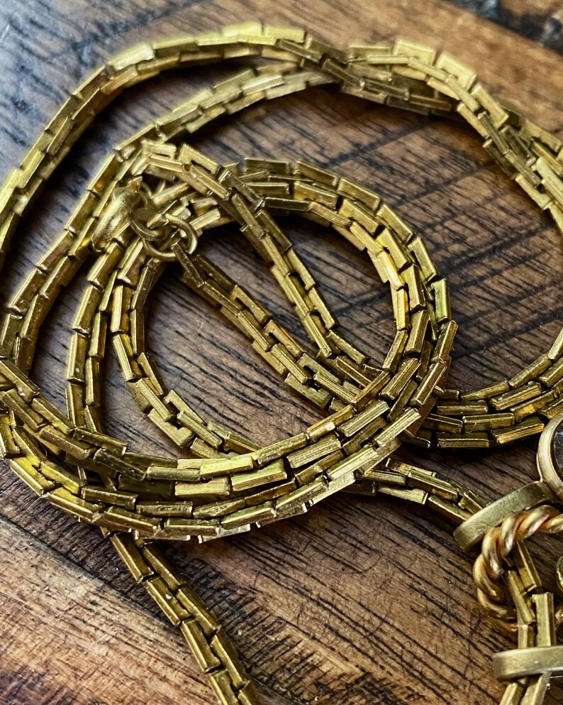 May include: A gold chain necklace with a box chain design. The chain is made of small, rectangular links.