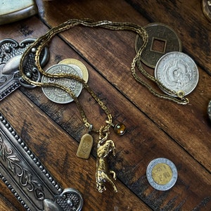 May include: A gold chain necklace with a tiger pendant, a small skull charm, and a brown gemstone. The necklace is laying on a wooden surface with several coins.