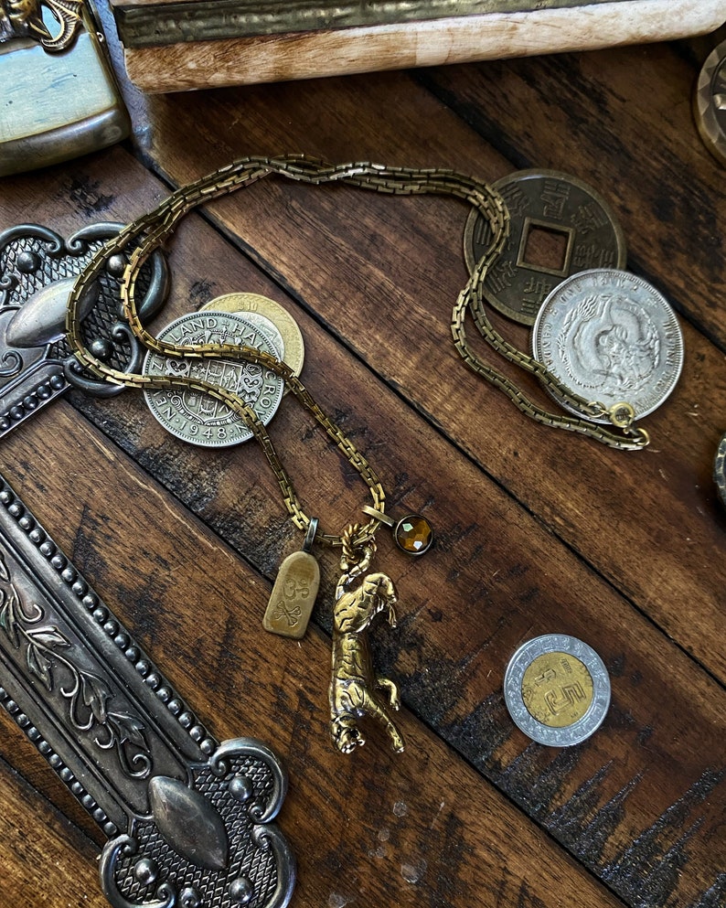 May include: A gold chain necklace with a tiger pendant, a small skull charm, and a brown gemstone. The necklace is laying on a wooden surface with several coins.