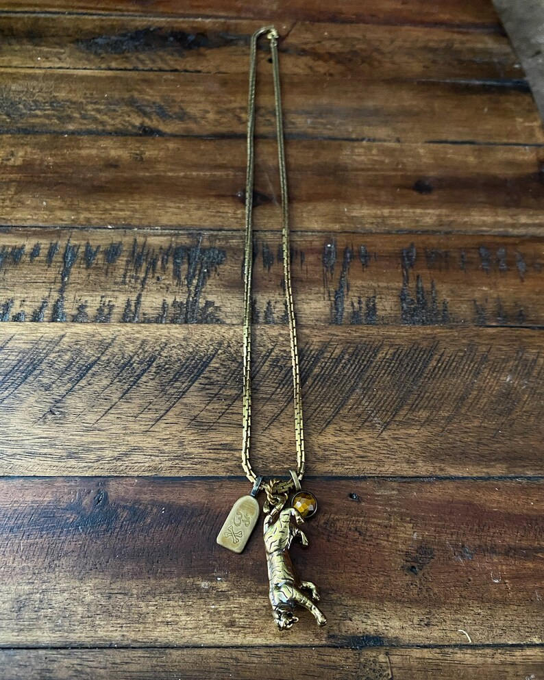 May include: A gold chain necklace with a tiger charm and a small amber bead. The tiger is holding a ball in its paws.