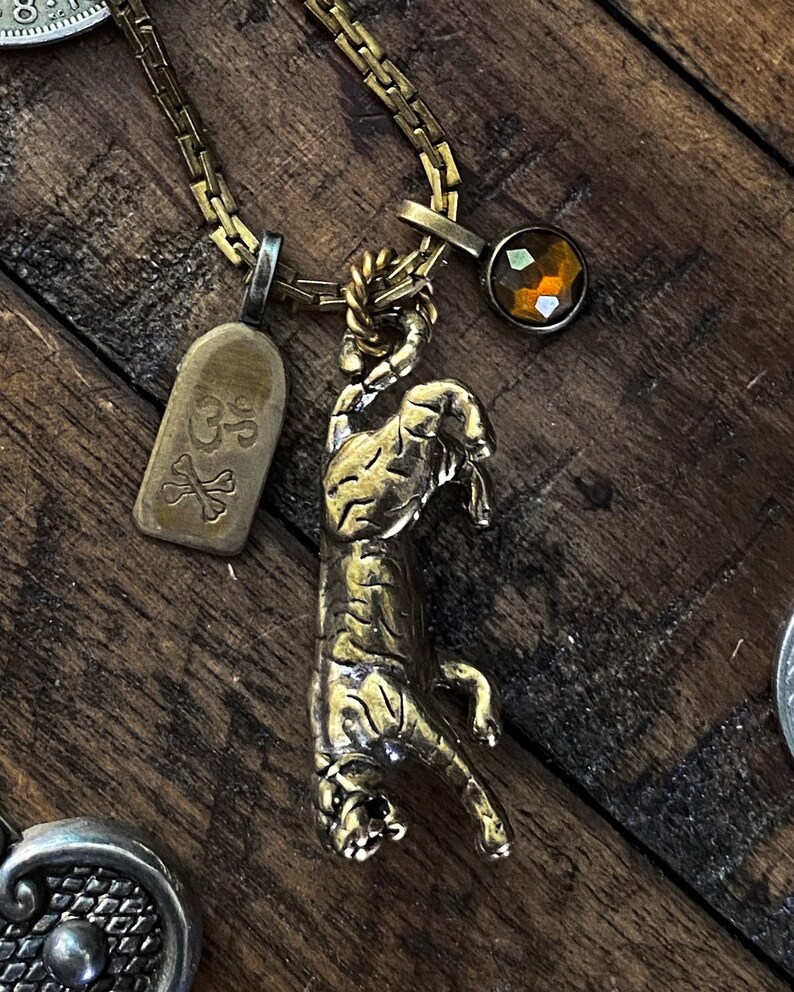 May include: A gold chain necklace with a tiger pendant, a small tag with the text "GP" and a crossbones design, and a brown faceted gemstone.