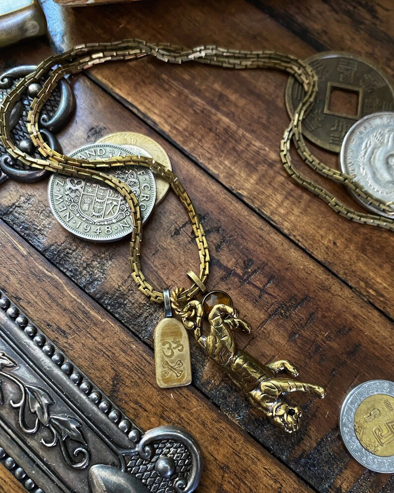 May include: A gold chain necklace with a tiger pendant and a small charm with the symbol "Om" on it. The necklace is lying on a wooden surface with several coins.