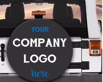 May include: A black spare tire cover with white text that says "YOUR COMPANY LOGO here" in a white Jeep Wrangler. The tire cover is on the back of the Jeep.