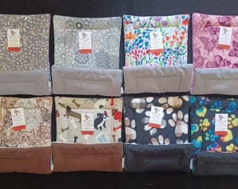 May include: Eight square fabric pet mats arranged in two rows. Each mat features a unique pattern including flowers, butterflies, cats, dogs, and paw prints. Each mat has a solid-colored rectangular bolster attached to the front edge.