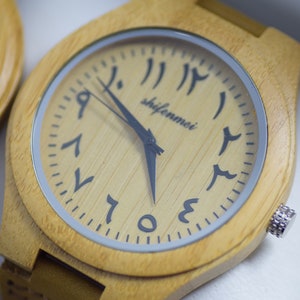 May include: A light-colored wooden wristwatch with Arabic numerals on the face. The watch has a square face with rounded corners and a silver-colored bezel. The watch hands are black, and the watch band is also light-colored wood.