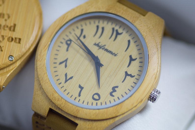 May include: A light-colored wooden wristwatch with Arabic numerals on the face. The watch has a square face with rounded corners and a silver-colored bezel. The watch hands are black, and the watch band is also light-colored wood.