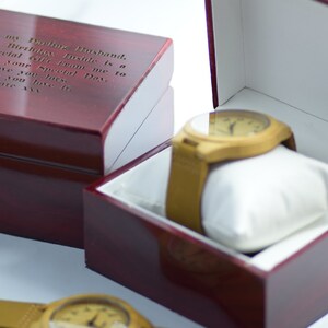 May include: A wooden watch with a tan leather strap rests on a white pillow inside a red wooden box. The box is open, revealing the watch. A second closed box with engraved text is next to it. The text reads: "To my Darling Husband..."