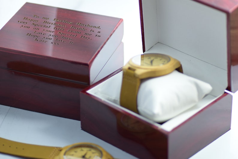 May include: A wooden watch with a tan leather strap rests on a white pillow inside a red wooden box. The box is open, revealing the watch. A second closed box with engraved text is next to it. The text reads: "To my Darling Husband..."