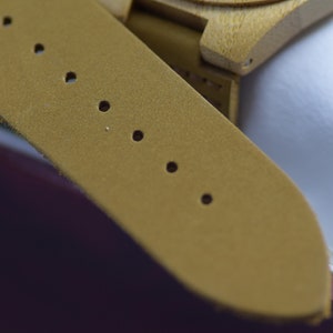 May include: Close-up of a mustard-colored watch strap with several evenly spaced holes. The strap is made of a textured material and is attached to a wooden watch face. The watch appears to be new.