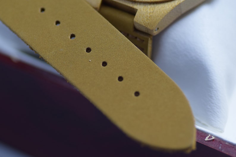 May include: Close-up of a mustard-colored watch strap with several evenly spaced holes. The strap is made of a textured material and is attached to a wooden watch face. The watch appears to be new.