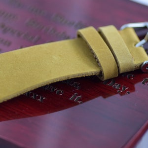 May include: Close-up of a tan leather watch strap with a silver buckle. The strap is made of a soft, textured leather and is attached to a polished silver buckle. The strap is resting on a dark red wooden surface.