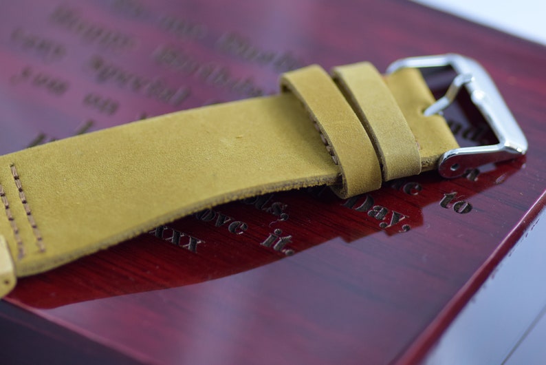 May include: Close-up of a tan leather watch strap with a silver buckle. The strap is made of a soft, textured leather and is attached to a polished silver buckle. The strap is resting on a dark red wooden surface.