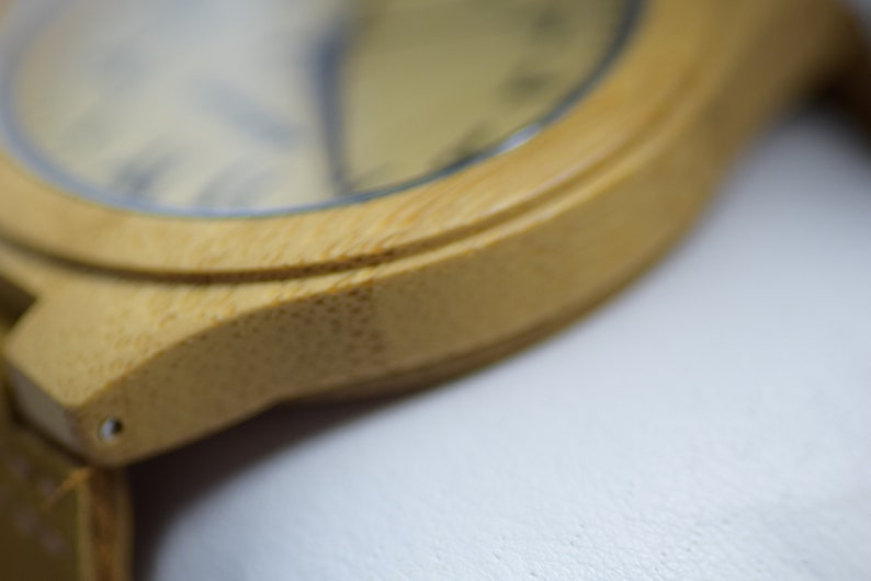 May include: Close-up of a bamboo watch. The watch has a light brown wooden frame and a light-colored watch face. The watch band is also made of bamboo. The watch is a natural, eco-friendly accessory.