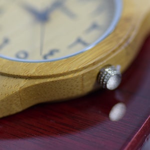 May include: Close-up of a wooden wristwatch with a light-colored face and Arabic numerals. The watch has a light brown leather strap and a silver crown. The watch is set against a dark red surface.