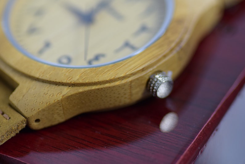 May include: Close-up of a wooden wristwatch with a light-colored face and Arabic numerals. The watch has a light brown leather strap and a silver crown. The watch is set against a dark red surface.