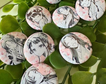 Op de afbeelding: Een verzameling ronde, glanzende knopen met anime-achtige illustraties van een vrouw met lang haar. De knopen hebben een roze bloemenachtergrond en zijn gerangschikt op een bed van groene bladeren.
