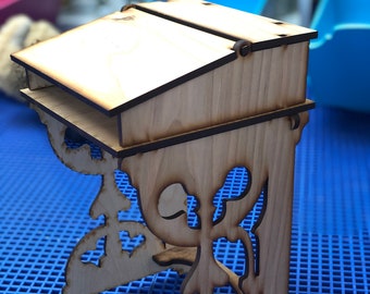 May include: A wooden desk with intricate cutouts on the legs. The desk has a hinged top and a shelf underneath. The desk is made of light brown wood and is sitting on a blue grid.