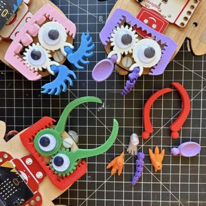 May include: A collection of colorful, 3D-printed robot parts and accessories, including eyes, gears, and articulated appendages. The parts are in various colors, such as pink, purple, green, and orange, and are arranged on a black grid surface.