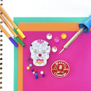 May include: A craft project setup featuring a decorated skull, colorful gems, and a soldering iron on a pink background. The skull is adorned with gems and gears. A red and white Maker Challenge badge is also present, along with colorful pens and paper.