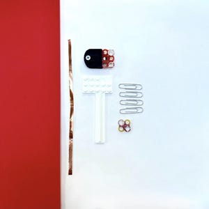 May include: An overhead shot of various small objects on a white surface, including a strip of copper tape, a black and red component, a white plastic piece, paper clips, and small colorful rings. A red background is on the left.