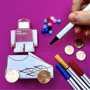 Puede incluir: Un fondo rosa presenta un robot de manualidades, un coche de papel, centavos, pompones de colores y rotuladores. El robot tiene el texto "little engineer". Una mano sostiene un bolígrafo blanco. El coche tiene diseños de llamas.