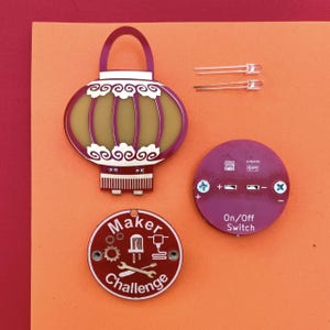 May include: A DIY maker kit with a purple and gold lantern, a red circular piece with the text "Maker Challenge", a purple circular piece with "On/Off Switch", and two LED lights, all on an orange background.