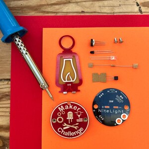 Może przedstawiać: Asortyment elementów elektronicznych do projektu DIY, w tym lutownica, czerwony i żółty akrylowy element w kształcie latarni oraz okrągła płytka drukowana z napisem "NiteLight Sensor". W zestawie znajduje się również czerwona i biała odznaka "Maker Challenge".
