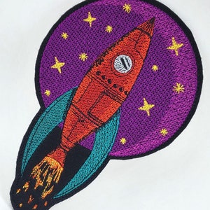 May include: Embroidered iron-on patch featuring a red rocket ship flying through a purple starry sky. The rocket has a teal flame and is surrounded by a teal and black circle.