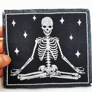 May include: Square black patch with a white skeleton in a yoga pose, surrounded by white diamond stars. The patch has a white stitched border.