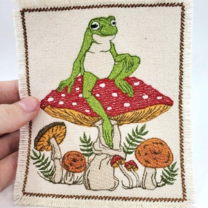 May include: Embroidered patch showing a green frog seated on a red mushroom with white spots. The patch has a cream background with brown stitching and a frayed edge. Other mushrooms and foliage are embroidered below.