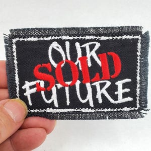May include: A rectangular black patch with white embroidered text that reads "OUR SOLD FUTURE" in a handwritten style. The word "SOLD" is in red. The patch has a white border and frayed edges.