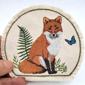 Może przedstawiać: Haftowana naszywka przedstawiająca lisa z pomarańczowym i białym futrem, zieloną paprocią i niebieskim motylem. Naszywka ma kremowe tło i postrzępioną krawędź.