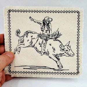 May include: Square, off-white fabric coaster with a black and white illustration of a rodeo cowboy riding a bull. The image is outlined with a black stitched border and has a frayed edge.