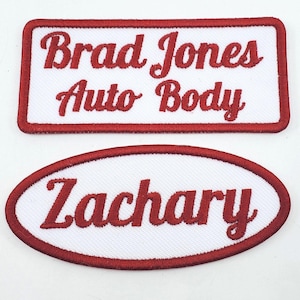 May include: Two embroidered patches with white fabric backgrounds and red borders. The rectangular patch features the red text Brad Jones Auto Body. The oval patch features the red text Zachary. Both patches have a stitched texture throughout.