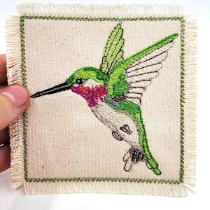 Könnte beinhalten: Gestickter Aufnäher mit einem grünen Kolibri mit rosa Kehle auf weißem Canvas-Hintergrund mit grünem Rand.