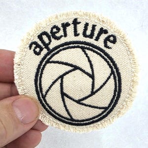 May include: A circular off-white fabric patch with a frayed edge featuring black embroidery. The design includes a camera aperture symbol in the center and the word aperture embroidered in black lowercase letters along the top curve.