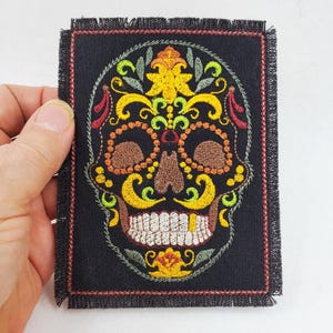 May include: A rectangular black fabric patch featuring an embroidered sugar skull design. The skull is decorated with intricate yellow, orange, green, and red floral patterns, brown eye sockets, and white teeth with a single gold tooth detail.