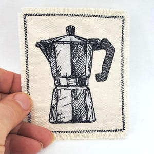Op de afbeelding: Een rechthoekige patch met een zwart-wit illustratie van een espressopot. De afbeelding is gedrukt op een beige stof met een gerafelde rand en zwarte stiksels. De patch wordt door een hand vastgehouden.
