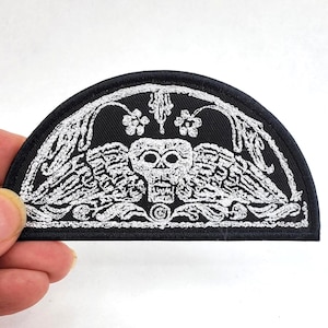 May include: A semi-circular embroidered patch featuring a black background with a detailed silver thread design. The artwork depicts a central skull with wings, surrounded by intricate floral and vine motifs, all enclosed within a solid black border.