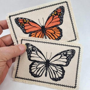 May include: Two butterfly patches. One patch features an orange and black monarch butterfly design. The other patch has a black and white butterfly design. Both patches are on a beige fabric with a black stitched border.
