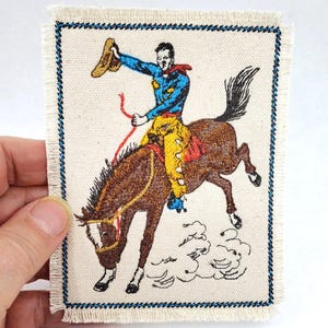 May include: A rectangular fabric patch with a frayed edge featuring an embroidered scene of a rider in a blue shirt and yellow chaps on a bucking brown horse. The rider holds a tan hat aloft, and the design is framed by a dark blue stitched border.