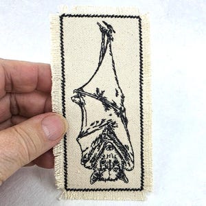 May include: A rectangular off-white fabric patch with frayed edges featuring a black ink illustration of a bat hanging upside down. The design is enclosed within a thin black rectangular border stitched onto the textured canvas material.