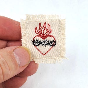 Puede incluir: Un pequeño parche cuadrado de tela color crema con bordes deshilachados que presenta un diseño bordado de un corazón rojo coronado por llamas y rodeado por una corona de espinas negra. El bordado está centrado en el cuadrado de tela.