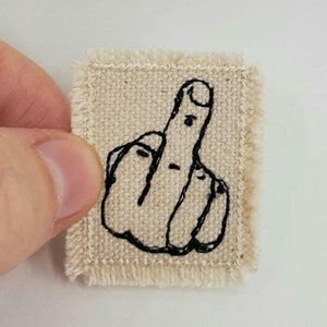 May include: A small, beige fabric patch with a black embroidered middle finger.