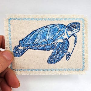 Pode incluir: Um remendo azul bordado de tartaruga marinha com fundo branco e borda azul claro. O remendo é costurado em uma tela branca.
