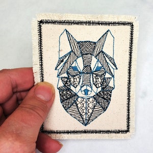 May include: A white canvas patch with a black stitched border. The patch features a geometric design of a wolf's head in black and blue thread.