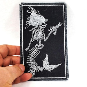 May include: A black embroidered patch with a white skeleton mermaid holding a fish. The mermaid has long flowing hair and a tail with a fin.