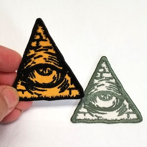 Può includere: Due toppe ricamate da stirare che rappresentano l'occhio che tutto vede in un triangolo. La toppa a sinistra è gialla con un occhio nero, e la toppa a destra è verde con un occhio bianco.